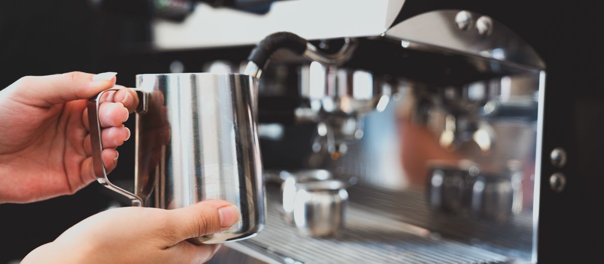 A barista steaming milk in a pitcher at an espresso machine.