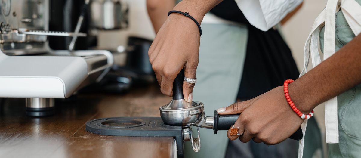 Side view crop faceless ethnic barista wearing apron pressing ground coffee by using tamper before preparing aromatic cappuccino while working in coffeehouse